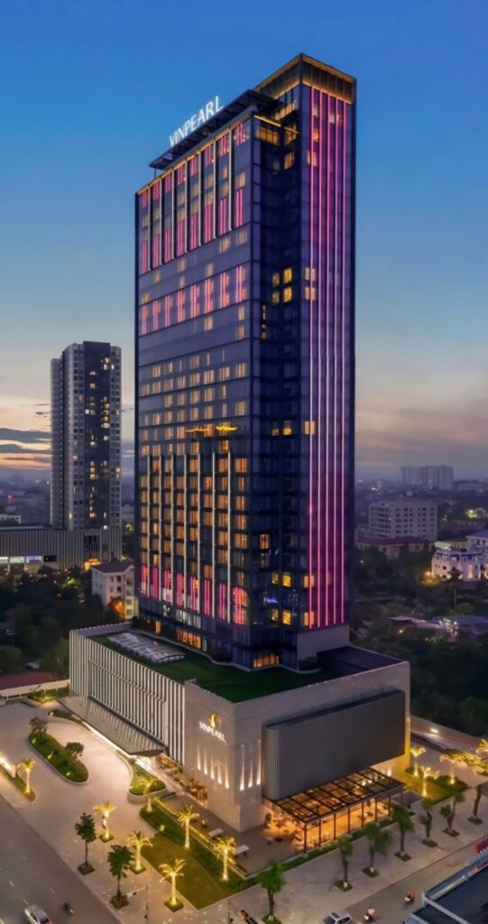 A tall, modern skyscraper with the name Vinpearl at the top, illuminated with vertical pink and purple lights at dusk, stands among other buildings in a cityscape.