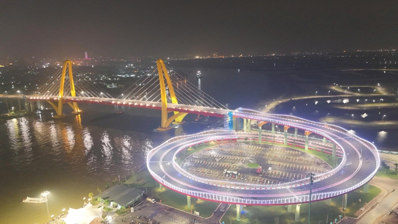 A brightly lit cable-stayed bridge with yellow towers spans a river at night. In the foreground, a circular, illuminated road structure curves over a parking area near the riverbank. City lights are visible in the background.